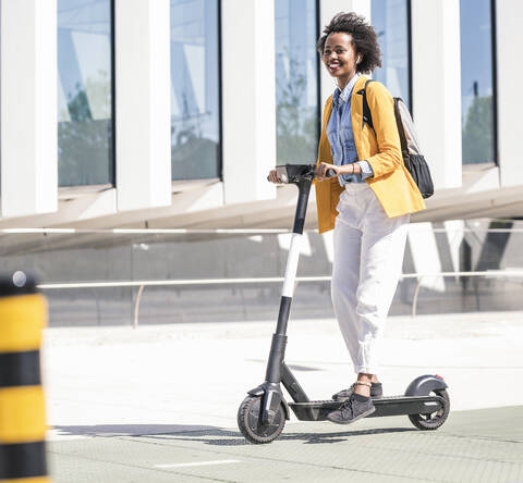 Glückliche junge Frau mit Kopfhörern auf einem E-Scooter in der Stadt, lizenzfreies Stockfoto