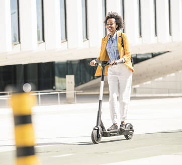 Happy young woman with earphones riding e-scooter in the city - UUF19622