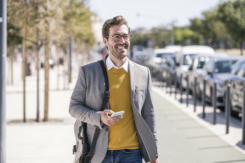 Lächelnder junger Geschäftsmann mit Mobiltelefon in der Stadt unterwegs, lizenzfreies Stockfoto