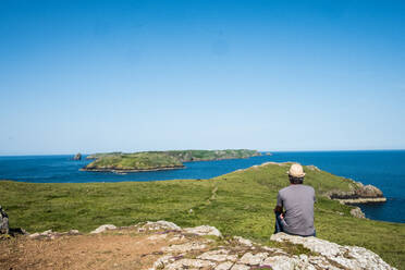 Rear view of man sitting on a cliff overlooking the Pembrokeshire Coast, Wales, UK. - MINF13038