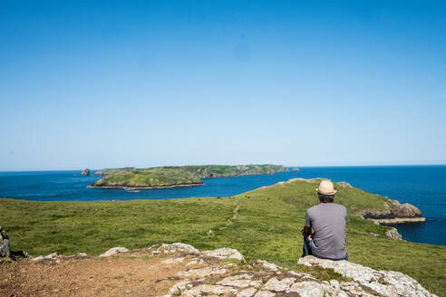 Rückansicht eines Mannes, der auf einer Klippe sitzt und die Pembrokeshire-Küste, Wales, Großbritannien, überblickt. - MINF13038