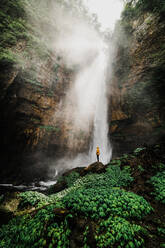 Male tourist in Indonesia looking at waterfall while sitting on rocks - CAVF68583