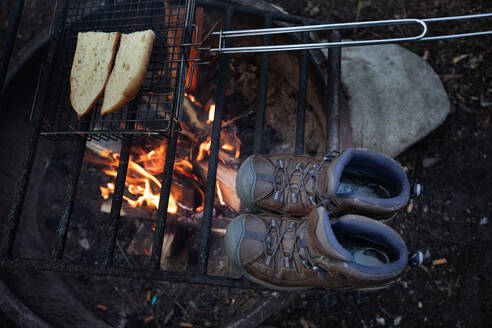 Nasse Wanderschuhe, die am Lagerfeuer trocknen, während Brot über den Flammen geröstet wird. - CAVF68622