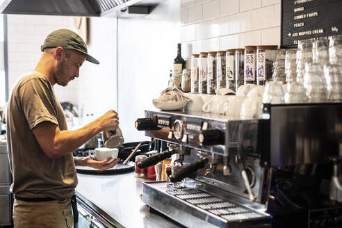 Bärtiger Mann mit Baseballmütze steht an einer Espressomaschine in einem Restaurant. - MINF13162