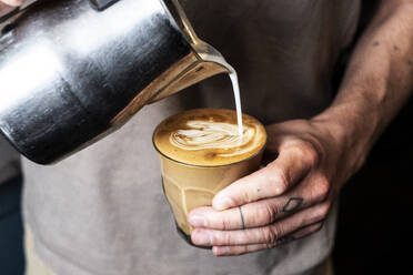 Nahaufnahme einer Person mit tätowiertem Finger, die Milch aus einer Metallkanne in ein Café-Latte-Glas gießt. - MINF13175