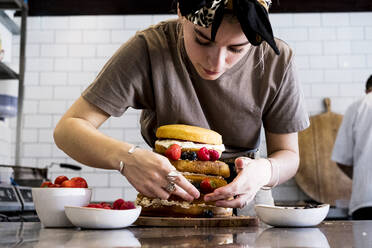 Ein Koch, der in einer Großküche arbeitet und einen geschichteten Biskuitkuchen mit frischen Früchten zusammenstellt. - MINF13206