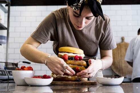A cook working in a commercial kitchen assembling a layered sponge cake with fresh fruit. - MINF13206