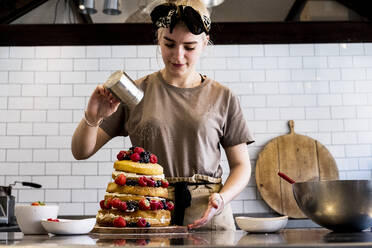 Ein Koch, der in einer Großküche arbeitet und Puderzucker über einen Schichtkuchen mit frischen Früchten streut. - MINF13210