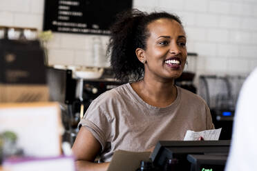 Eine Barista-Frau lächelt zur Begrüßung neben dem Tresen eines Coffeeshops. - MINF13212
