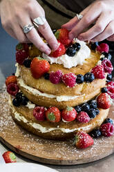 Eine Köchin, die eine Torte mit frischer Sahne und frischem Obst, Erdbeeren und Blaubeeren, zusammenstellt. - MINF13216