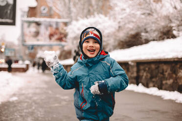 Glücklicher Junge hält Schneeball beim Laufen auf der Straße im Winter - CAVF68784