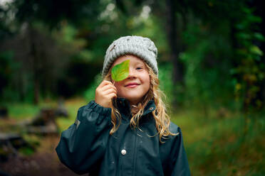 Lustiges Mädchen in warmer Mütze schließt die Augen mit grünem Blatt im Wald - CAVF68820