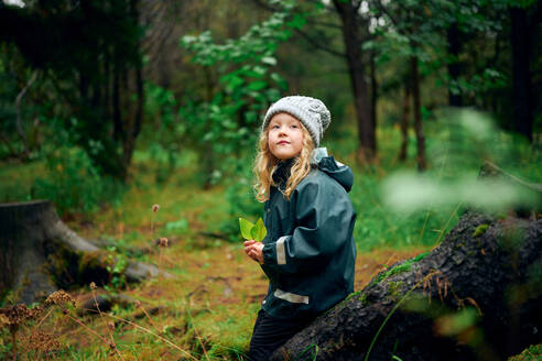 Small girl in hat and jacket sitting on tree stump and looking up - CAVF68821