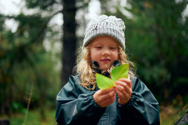 Pretty blonde girl in warm hat holding green leaves in hand - CAVF68822