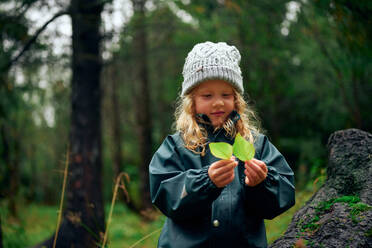 Curly girl in gray warm hat holding green leaves in forest - CAVF68823