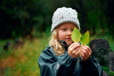 Charming girl in warm hat holding green leaves in forest - CAVF68826