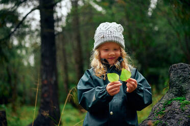 Charmantes Mädchen in warmer Mütze hält grüne Blätter im Wald - CAVF68832