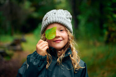Long haired charming girl in warm hat closing eye with green leaf in forest - CAVF68834