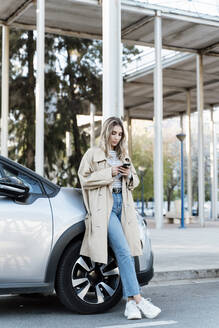 Junge blonde Frau, die ein Smartphone benutzt und sich an ein Auto lehnt - ERRF02027