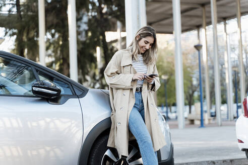 Junge blonde Frau, die ein Smartphone benutzt und sich an ein Auto lehnt - ERRF02028