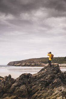 Young woman wearing yellow rain jacket at the beach, looking through binoculars, Bretagne, France - UUF19645