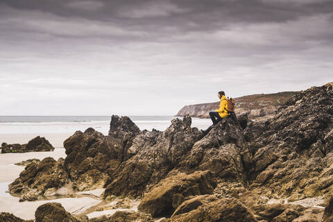 Junge Frau in gelber Regenjacke sitzt auf einem Felsen am Strand, Bretagne, Frankreich, lizenzfreies Stockfoto