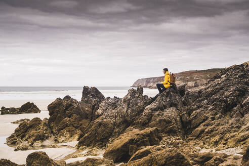 Junge Frau in gelber Regenjacke sitzt auf einem Felsen am Strand, Bretagne, Frankreich - UUF19647