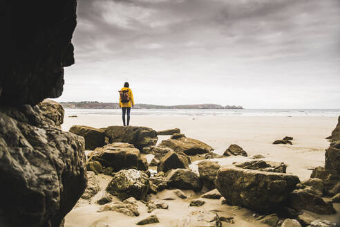 Young woman wearing yellow rain jacket at the beach at rock cave, Bretagne, France - UUF19649