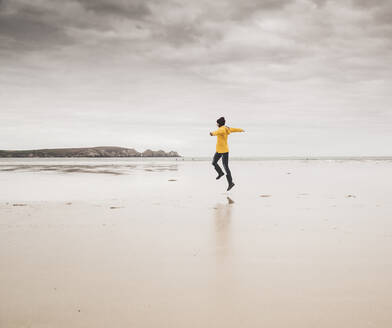 Young woman wearing yellow rain jacket, jumping at the beach, Bretagne, France - UUF19654