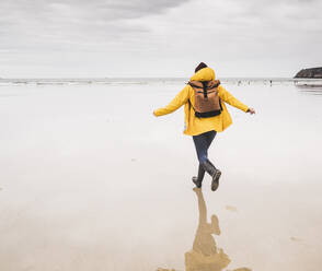 Junge Frau in gelber Regenjacke und beim Laufen am Strand, Bretagne, Frankreich - UUF19657