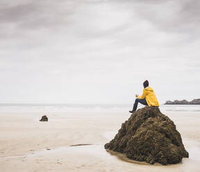 Junge Frau in gelber Regenjacke sitzt auf einem Felsen am Strand, Bretagne, Frankreich - UUF19662