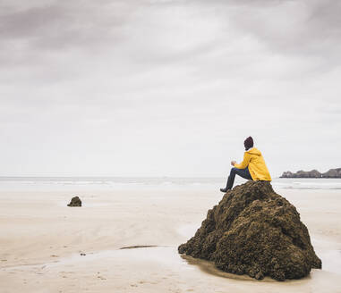 Young woman wearing yellow rain jacket sitting on rock at the beach, Bretagne, France - UUF19662