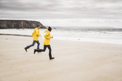 Junge Frau in gelben Regenjacken, die am Strand läuft, Bretagne, Frankreich, lizenzfreies Stockfoto