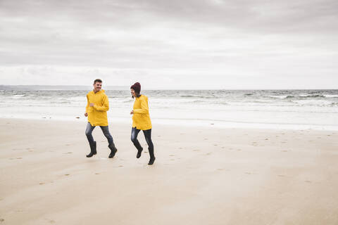 Junge Frau in gelben Regenjacken, die am Strand läuft, Bretagne, Frankreich, lizenzfreies Stockfoto