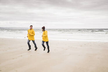 Young woman wearing yellow rain jackets and running at the beach, Bretagne, France - UUF19670