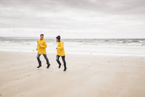 Young woman wearing yellow rain jackets and running at the beach, Bretagne, France - UUF19670