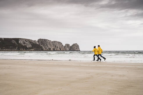 Young woman wearing yellow rain jackets and running at the beach, Bretagne, France - UUF19671