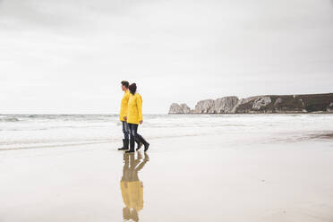 Young woman wearing yellow rain jackets and walking along the beach, Bretagne, France - UUF19672