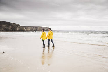 Young woman wearing yellow rain jackets and walking along the beach, Bretagne, France - UUF19674