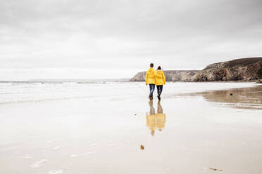 Young woman wearing yellow rain jackets and walking along the beach, Bretagne, France - UUF19675