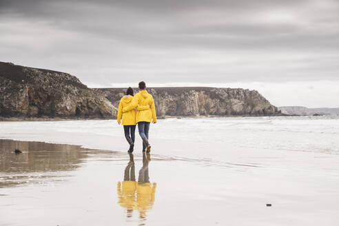 Young woman wearing yellow rain jackets and walking along the beach, Bretagne, France - UUF19676