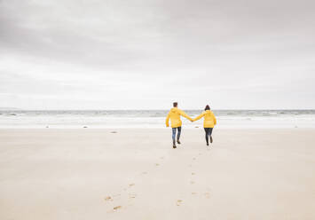 Junge Frau in gelben Regenjacken, die am Strand entlang läuft, Bretagne, Frankreich - UUF19678