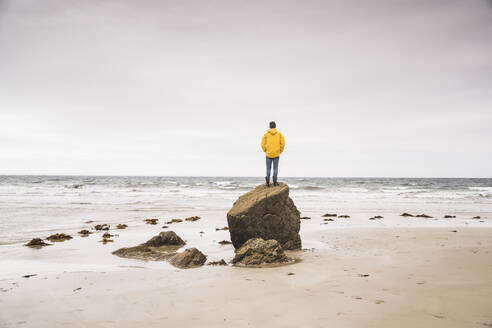 Junger Mann mit gelber Regenjacke am Strand und auf einem Felsen stehend, Bretagne, Frankreich - UUF19679