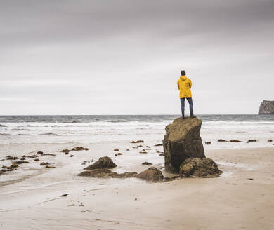 Junger Mann mit gelber Regenjacke am Strand und auf einem Felsen stehend, Bretagne, Frankreich - UUF19680