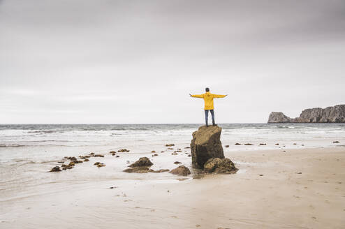 Junger Mann mit gelber Regenjacke am Strand und auf einem Felsen stehend, Bretagne, Frankreich - UUF19681