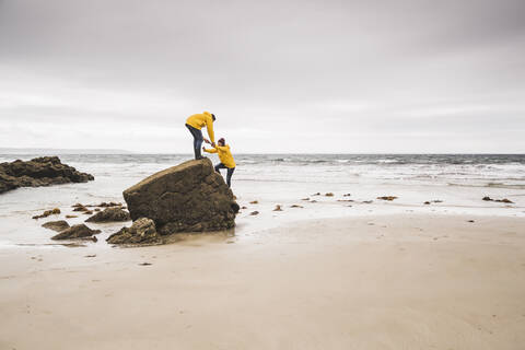 Junge Frau in gelber Regenjacke, die auf einen Felsen am Strand klettert, Bretagne, Frankreich, lizenzfreies Stockfoto