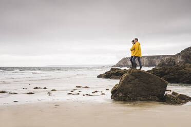 Young woman wearing yellow rain jackets and standing on rock at the beach, Bretagne, France - UUF19683