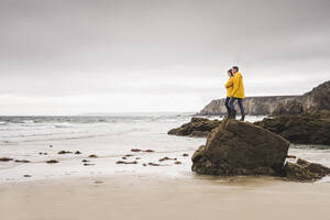 Junge Frau in gelben Regenjacken, die auf einem Felsen am Strand steht, Bretagne, Frankreich - UUF19683