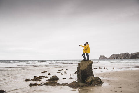 Junge Frau in gelben Regenjacken, die auf einem Felsen am Strand steht, Bretagne, Frankreich, lizenzfreies Stockfoto