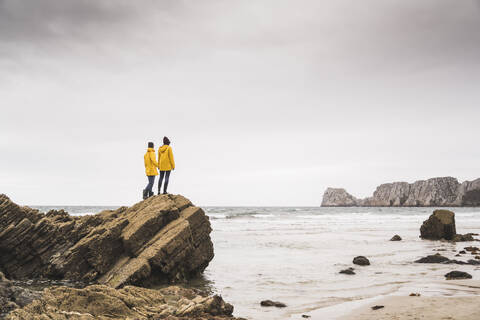 Young woman wearing yellow rain jackets and standing on rock at the beach, Bretagne, France stock photo
